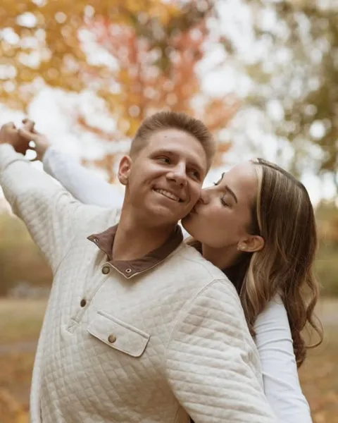A professional outdoor portrait of a smiling couple, with the woman kissing the man's cheek against a background of autumn foliage.