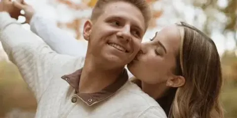 A professional outdoor portrait of a smiling couple, with the woman kissing the man's cheek against a background of autumn foliage.
