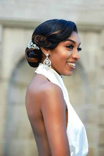A smiling woman in profile wearing a white dress, styled hair with a jeweled clip, and professional makeup.