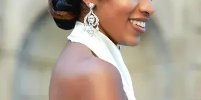 A smiling woman in profile wearing a white dress, styled hair with a jeweled clip, and professional makeup.