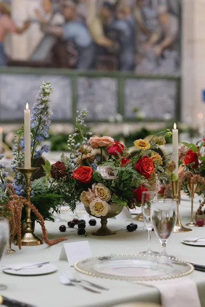 A lush, garden-style floral arrangement featuring red and peach roses, delphiniums, and greenery in a brass bowl on a wedding table setting.