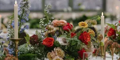 A lush, garden-style floral arrangement featuring red and peach roses, delphiniums, and greenery in a brass bowl on a wedding table setting.