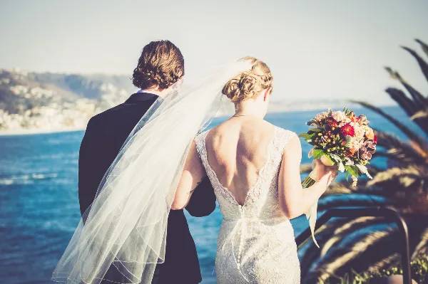 A bride and groom standing together looking out at a coastal landscape, with the bride holding a bouquet.