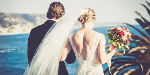 A bride and groom standing together looking out at a coastal landscape, with the bride holding a bouquet.