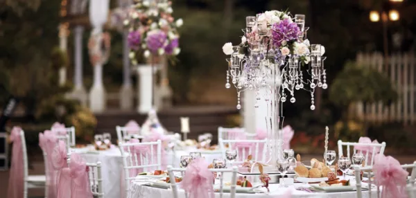 A catering table set with white linens, pink chair sashes, floral centerpieces, and plated food.