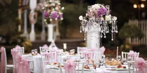 A catering table set with white linens, pink chair sashes, floral centerpieces, and plated food.