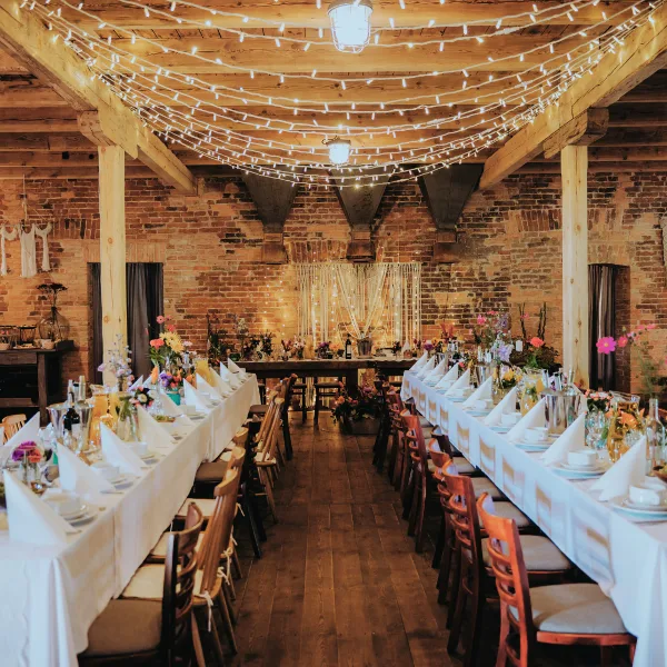 Wedding reception hall with long tables, wooden chairs, floral centerpieces, and overhead string lights.