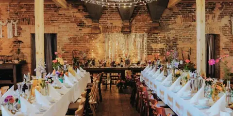 Wedding reception hall with long tables, wooden chairs, floral centerpieces, and overhead string lights.