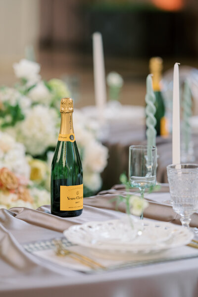 A close-up of an elegant wedding table setting featuring a bottle of Veuve Clicquot champagne, white floral arrangements, and crystal glassware.