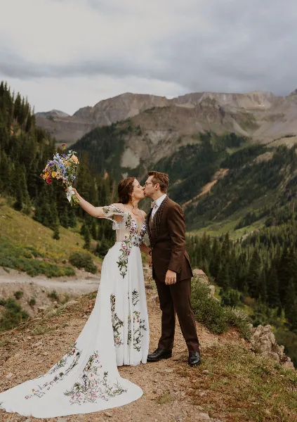 A bride and groom kissing on a mountain trail with a bouquet, set against a backdrop of pine trees and rugged peaks.