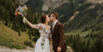 A bride and groom kissing on a mountain trail with a bouquet, set against a backdrop of pine trees and rugged peaks.