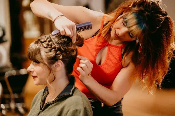 A hair stylist with blue-streaked hair styling a client's hair into a braided updo.