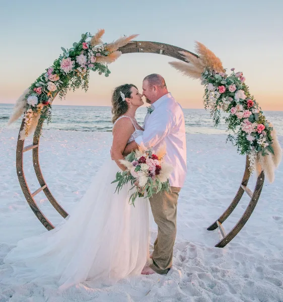 A bride and groom kissing under a floral arch on a beach at sunset.