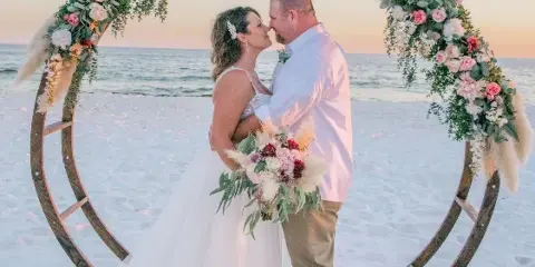 A bride and groom kissing under a floral arch on a beach at sunset.
