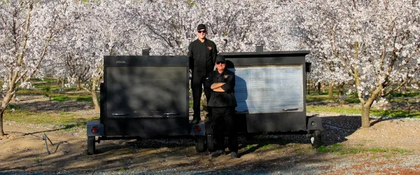 Two catering staff members standing in front of a mobile BBQ kitchen trailer in an orchard.