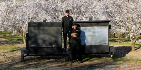 Two catering staff members standing in front of a mobile BBQ kitchen trailer in an orchard.