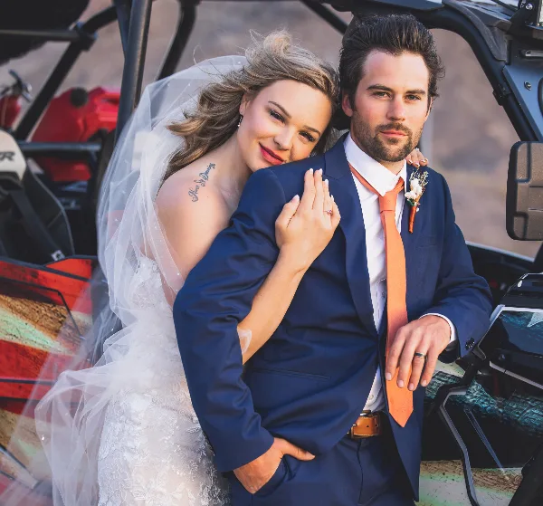 A bride and groom posing together in front of an off-road vehicle, with the bride wearing a white lace dress and veil and the groom in a blue suit with an orange tie.