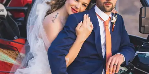 A bride and groom posing together in front of an off-road vehicle, with the bride wearing a white lace dress and veil and the groom in a blue suit with an orange tie.