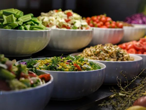 A catering buffet display featuring several large white bowls filled with various prepared salads, including a corn salad, coleslaw, and chopped vegetables.