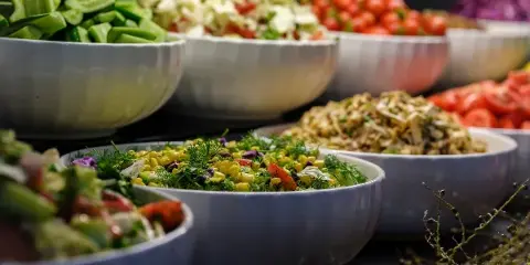 A catering buffet display featuring several large white bowls filled with various prepared salads, including a corn salad, coleslaw, and chopped vegetables.