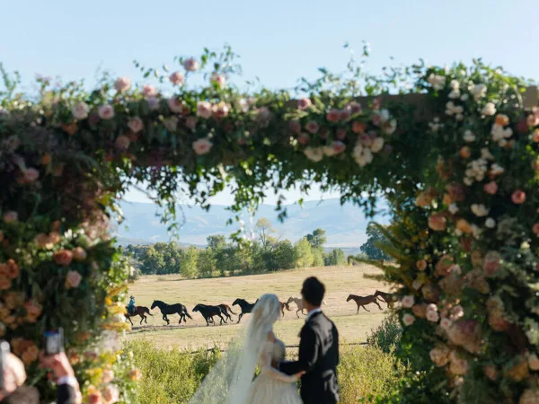 A bride and groom stand under a floral arch during a wedding ceremony with horses running in the background.