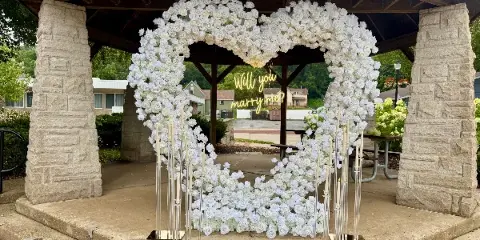 A large white floral heart arch with tall candles set up in a stone gazebo.