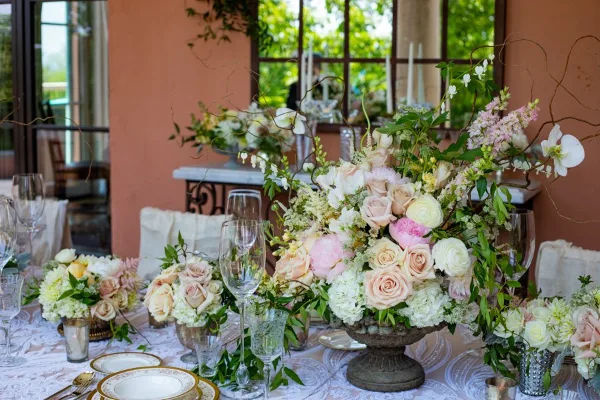 A wedding table setting featuring a large centerpiece of pink and white roses, hydrangeas, and greenery, accompanied by smaller floral arrangements and glassware.