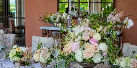 A wedding table setting featuring a large centerpiece of pink and white roses, hydrangeas, and greenery, accompanied by smaller floral arrangements and glassware.