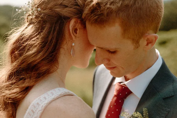 A close-up, candid photograph of a bride and groom in an outdoor setting, showing the couple in an intimate moment with soft, natural lighting.
