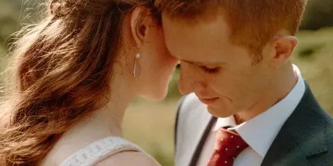A close-up, candid photograph of a bride and groom in an outdoor setting, showing the couple in an intimate moment with soft, natural lighting.