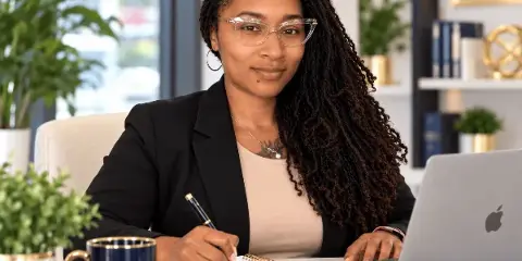 A professional woman in a business suit sitting at a desk with a laptop, notebook, and pen.