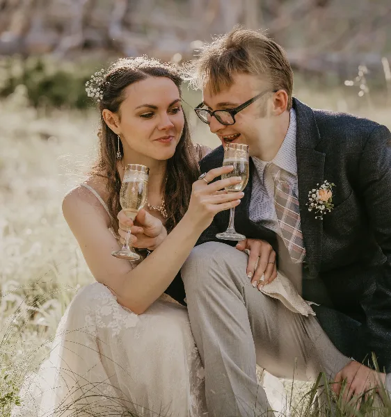 A bride and groom sitting in a grassy field, holding champagne flutes and smiling at each other.