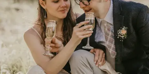 A bride and groom sitting in a grassy field, holding champagne flutes and smiling at each other.