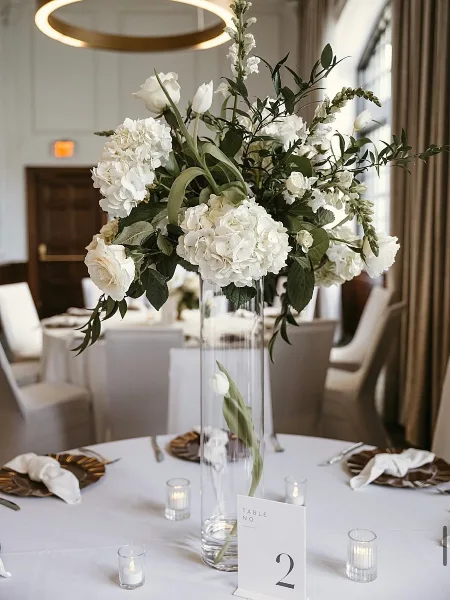 A tall glass vase filled with white hydrangeas and roses serves as a centerpiece on a white tablecloth, accompanied by gold-rimmed plates and a table number card.