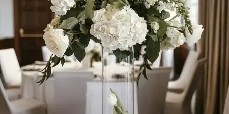 A tall glass vase filled with white hydrangeas and roses serves as a centerpiece on a white tablecloth, accompanied by gold-rimmed plates and a table number card.