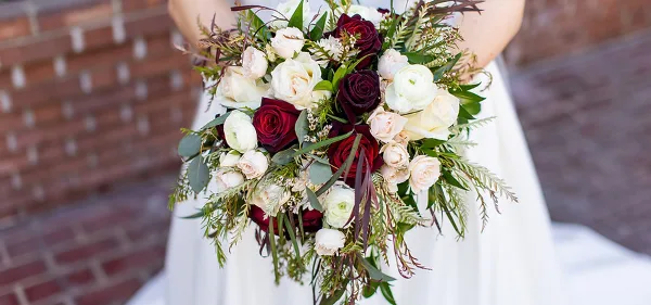 A bride holding a lush wedding bouquet featuring deep red and white roses, ranunculus, and greenery against a brick background.