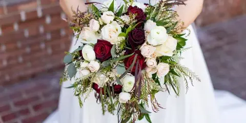 A bride holding a lush wedding bouquet featuring deep red and white roses, ranunculus, and greenery against a brick background.