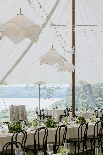 Elegant wedding reception tables with floral centerpieces and hanging lights inside a large white tent.