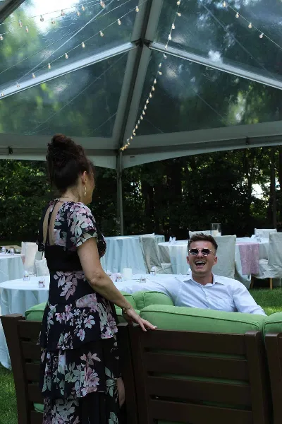 A candid photograph of a smiling couple interacting at a wedding reception under a clear tent with string lights.