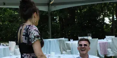 A candid photograph of a smiling couple interacting at a wedding reception under a clear tent with string lights.