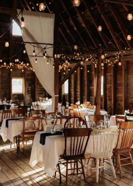 Interior of a historic barn wedding venue featuring wooden rafters, string lights, and reception tables set with white linens.