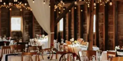 Interior of a historic barn wedding venue featuring wooden rafters, string lights, and reception tables set with white linens.