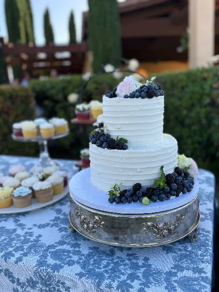 A two-tier white wedding cake decorated with blueberries and blackberries, displayed on a silver stand next to a tiered tray of floral cupcakes.