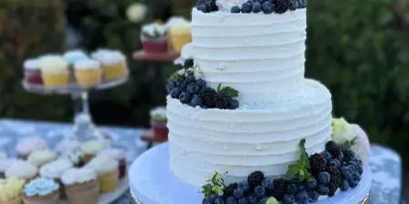 A two-tier white wedding cake decorated with blueberries and blackberries, displayed on a silver stand next to a tiered tray of floral cupcakes.