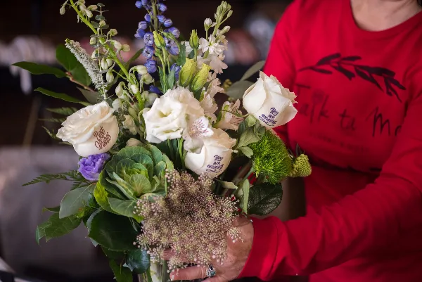 A person in a red 'Talk ta Me' shirt holding a custom bouquet of white roses with personalized messages embossed on the petals.