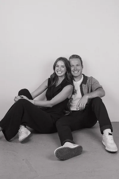 A black-and-white photograph of a smiling couple sitting on the floor against a plain white wall.