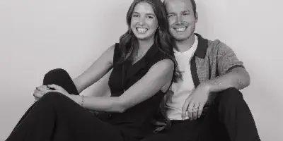 A black-and-white photograph of a smiling couple sitting on the floor against a plain white wall.
