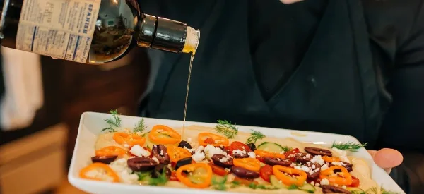 A chef in a black uniform pouring olive oil over a plated dish featuring sliced peppers, olives, and feta cheese.