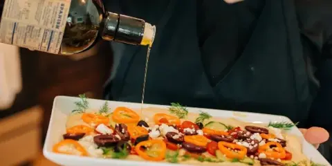 A chef in a black uniform pouring olive oil over a plated dish featuring sliced peppers, olives, and feta cheese.