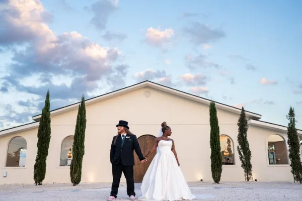 A bride and groom holding hands in front of a white building with tall cypress trees under a cloudy sky.